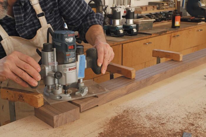 Tom routs the shoulder of the tenon on the stretcher with a router, using a pattern bit, cleaning up the surface left behind by the table saw cuts. The streatcher is secured to his benchtop using two clamps. 