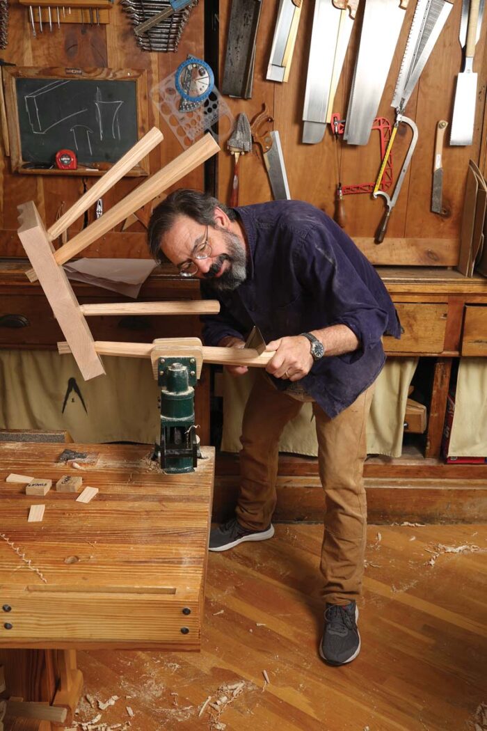 Schwarz is sawing the end of the chair leg off to create a chair that sits level. He is hunched down in a way that makes my back hurt to look at. 