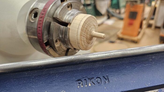 A piece of wood mounted in the chuck of a lathe. The wood has been worked to have a pattern cut into the face of it. 